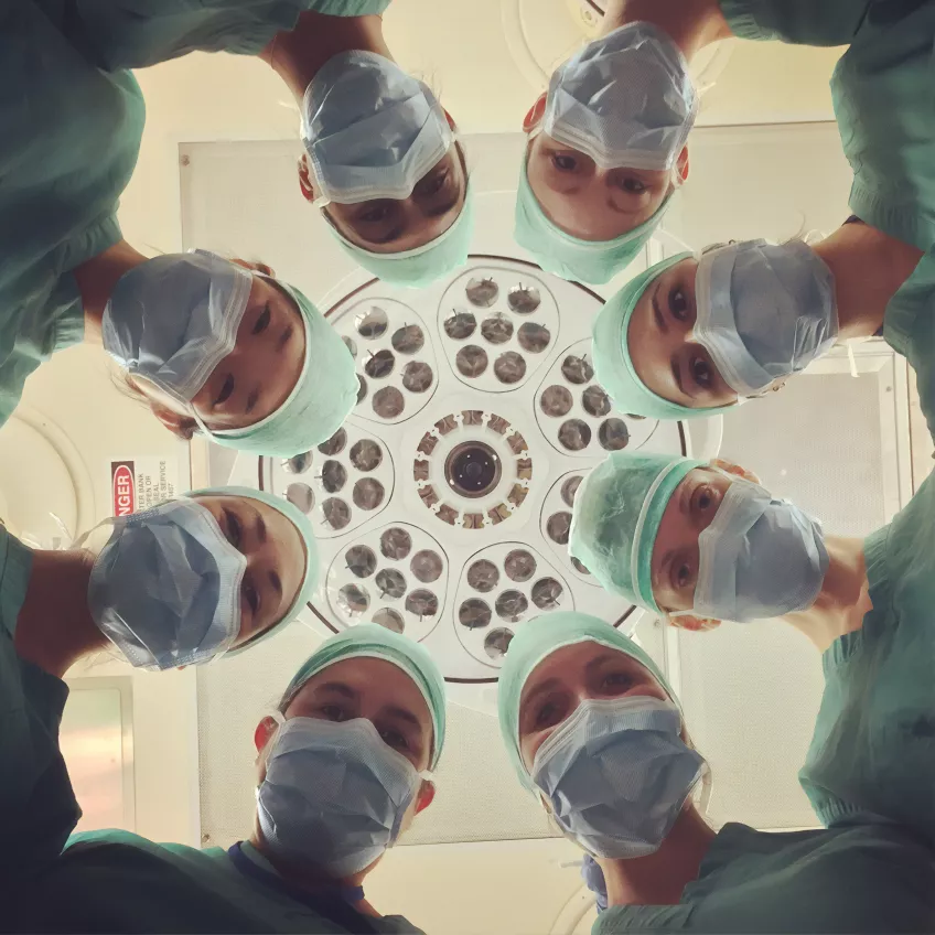 Several surgeons in scrubs and with face masks in a circle looking down. Photo.