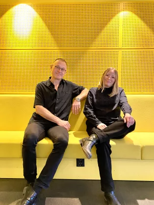 A man and a woman wearing black sitting in front of a yellow wall. Photo.